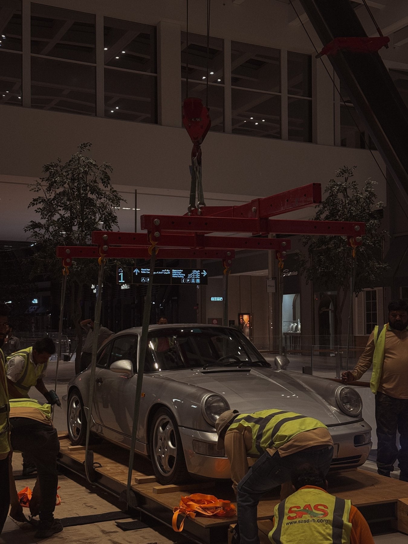 A silver Porsche 964 RS lifted by spider crane into Dubai Hills Mall at night